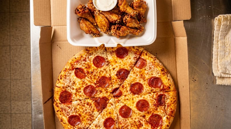 Pepperoni pizza in a cardboard box next to a container of chicken wings and dipping sauce.