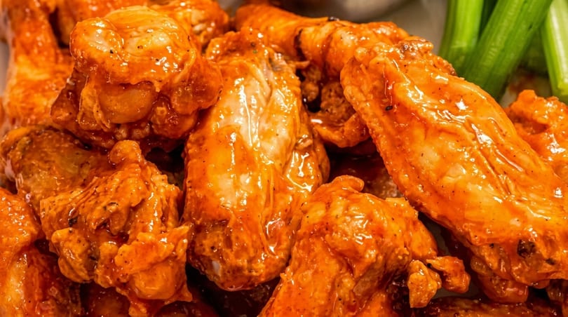 Close-up of buffalo chicken wings coated in orange sauce, with celery sticks in the background.
