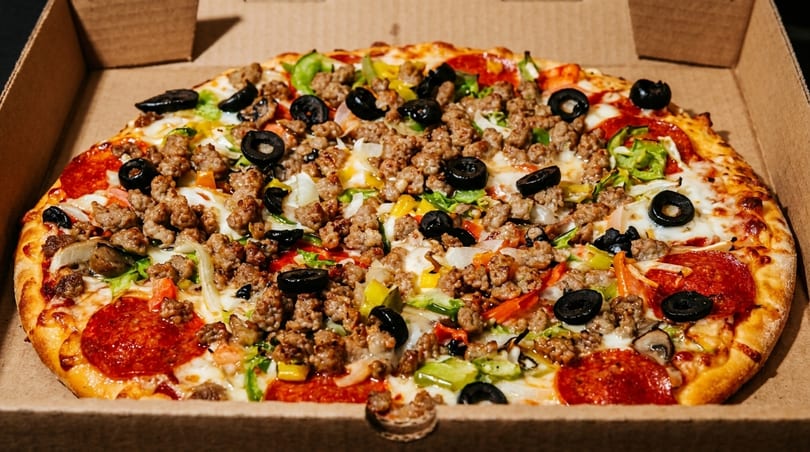 A whole pizza with pepperoni, sausage, olives, and vegetables in a cardboard box.